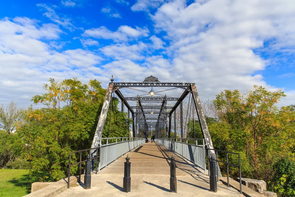 View of an old bridge in New Braunfels, Texas, one of the best places to visit in Texas, with tall green trees on either side of the walking path