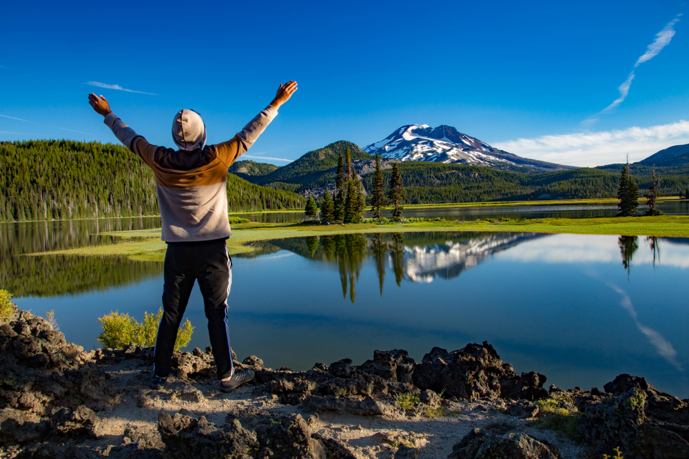 Man holding his arms above his head and standing on the edge of a lake while he looks at a mountain in the distance during the best time to visit Bend, Oregon