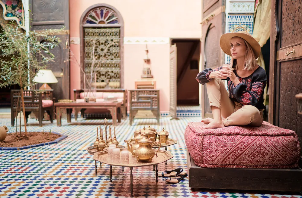 Thin blonde woman in a boho-style hat sitting cross-legged ion a red pillow drinking tea during the best time to go to Marrakech