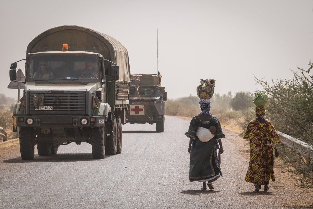 Women with baskets on their head walking by a convoy of military trucks with a sandy cover over the desert in the background to illustrate that Mali is not safe to visit right now
