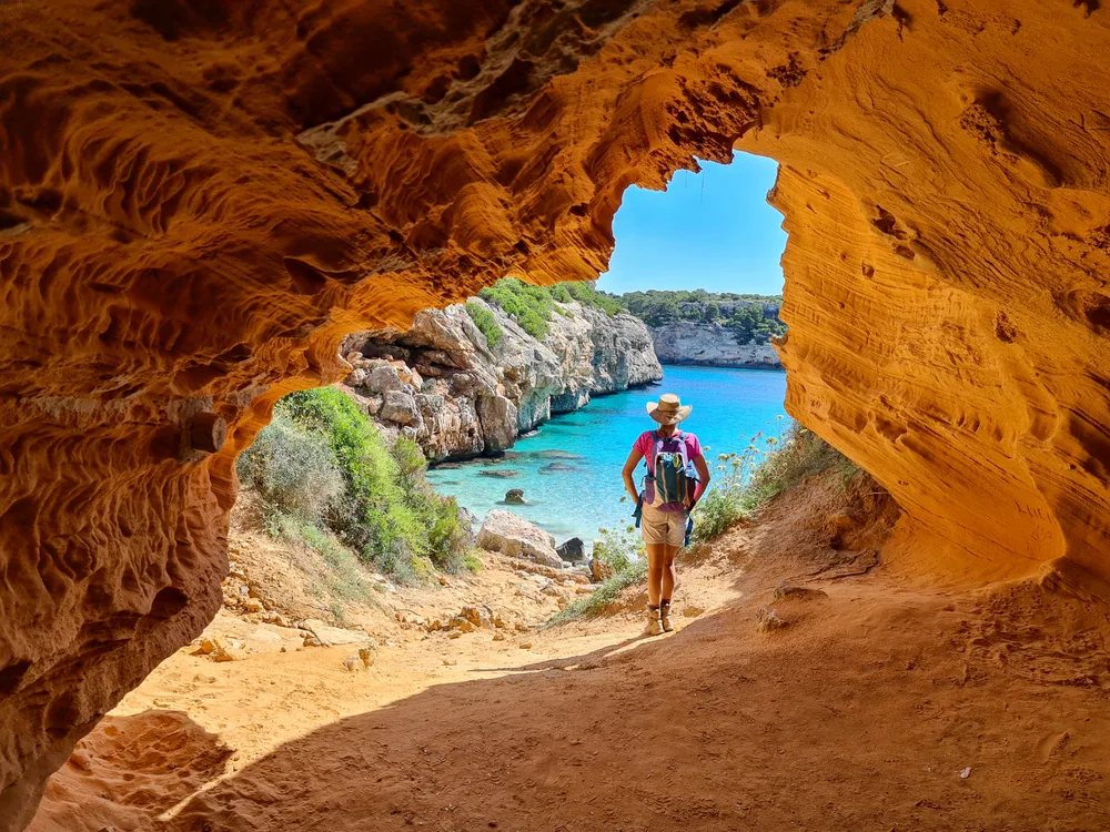 Thin tourist woman in a hiking hat and hiking shorts standing in the middle of a circular rock formation overlooking the deep blue water of the ocean during the best time to visit Mallorca