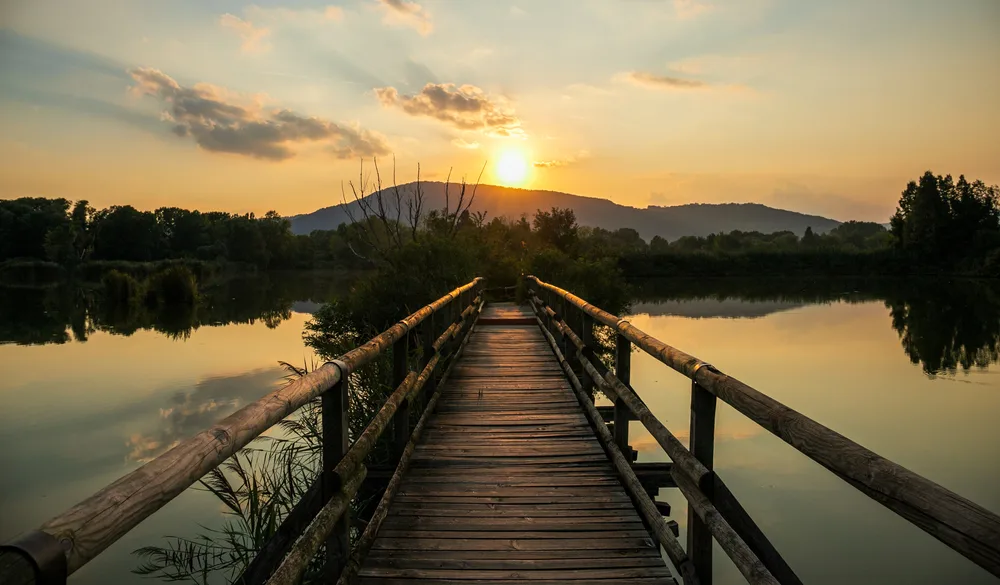 Wooden walking path with railings pictured at dusk with the sun setting over still water with mountains in the background for a piece on the least busy time to visit Italian Lakes