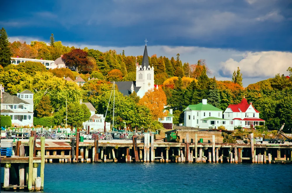 Autumn view of Mackinac Island, one of the best places to go to in September, as seen from a boat in the water with a church and some homes with unique colored roofs on the water's edge