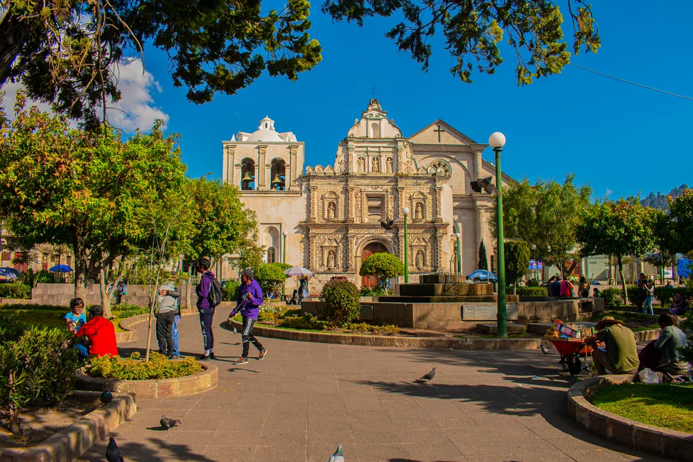 Xela in Quetzaltenango with the church pictured behind an idyllic garden and walking area with people mulling about on a blue-sky day for a piece on the must-visit places in Guatemala