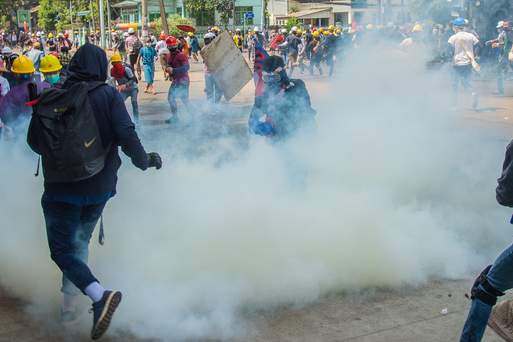 To show that Myanmar is not safe to visit, a person running through tear gas behind police with shields