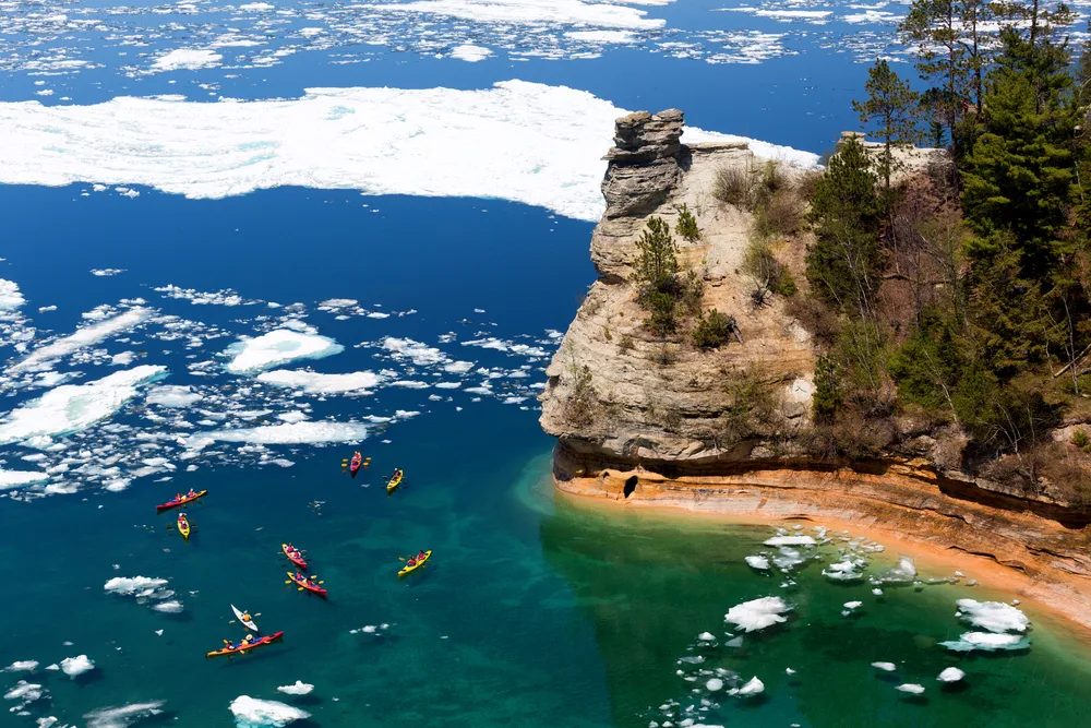 Kayaks go through ice formations next to a rocky cliff during the cheapest time to visit Pictured Rocks