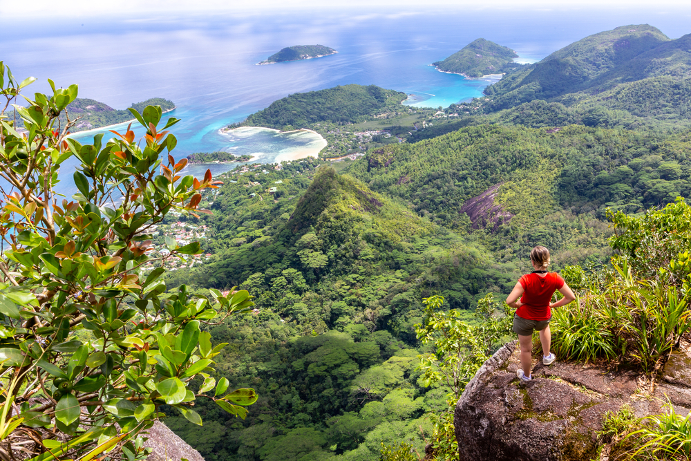 A female traveler stands on a cliff overlooking the island rain forest and ocean for a piece comparing adventure in Seychelles vs Maldives