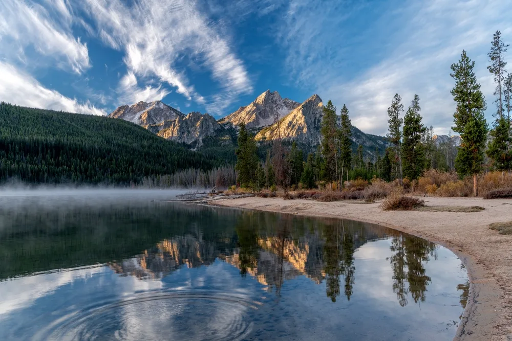 Sun rising over the Sawtooth Mountains with a still pond in the middle of some pine trees pictured during the best time to visit Idaho