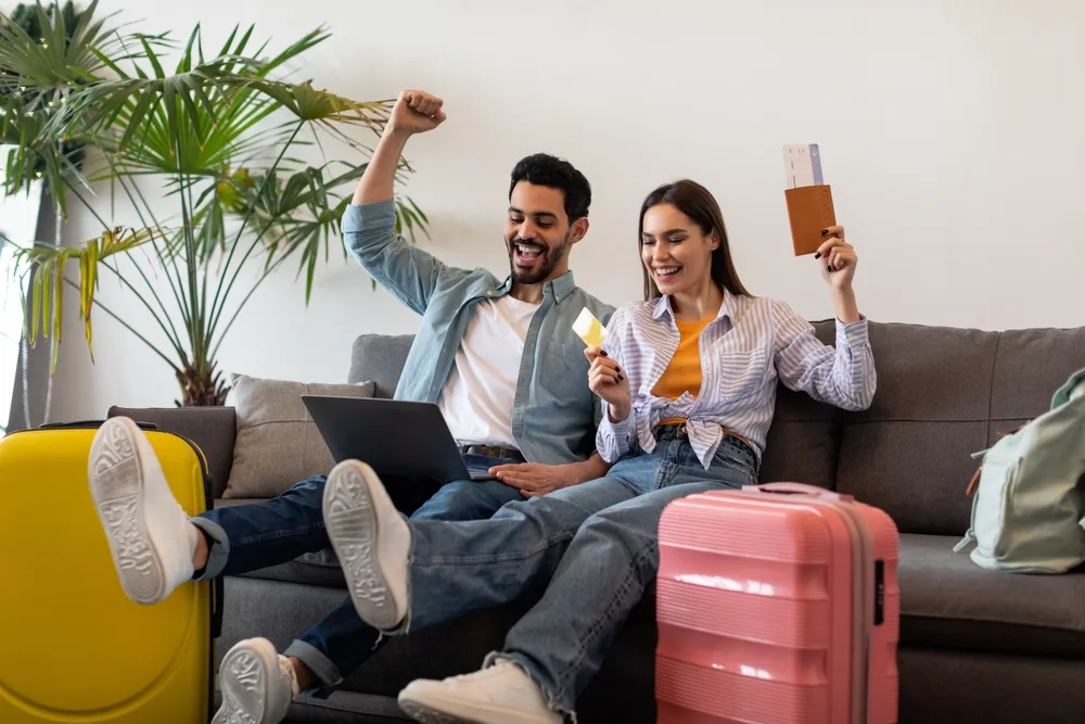 Couplec celebrates sitting on the couch with luggage packed after booking flight for a piece on how to find cheap flights