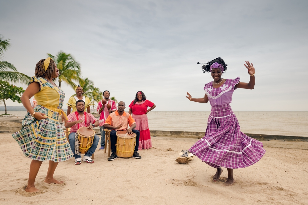 Pretty black woman in colorful dresses pictured dancing on a sandy beach in Honduras during the best time to visit with cloudy skies behind them
