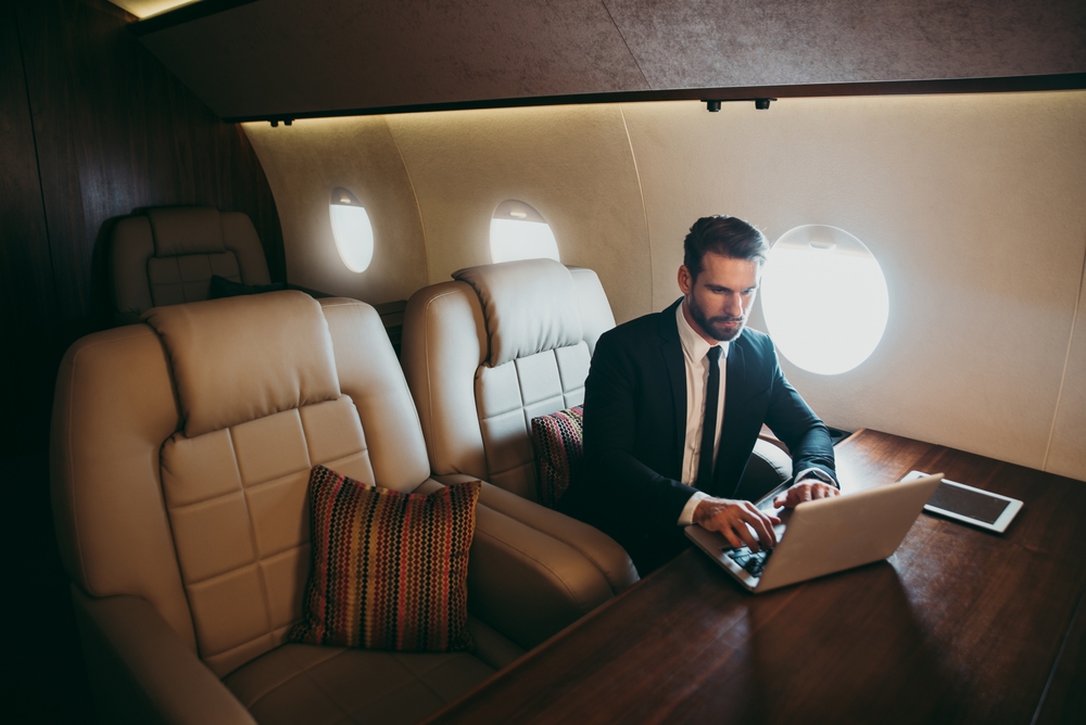 Businessman sits in a large private seat using his laptop on a plane for a comparison between business class vs. first class