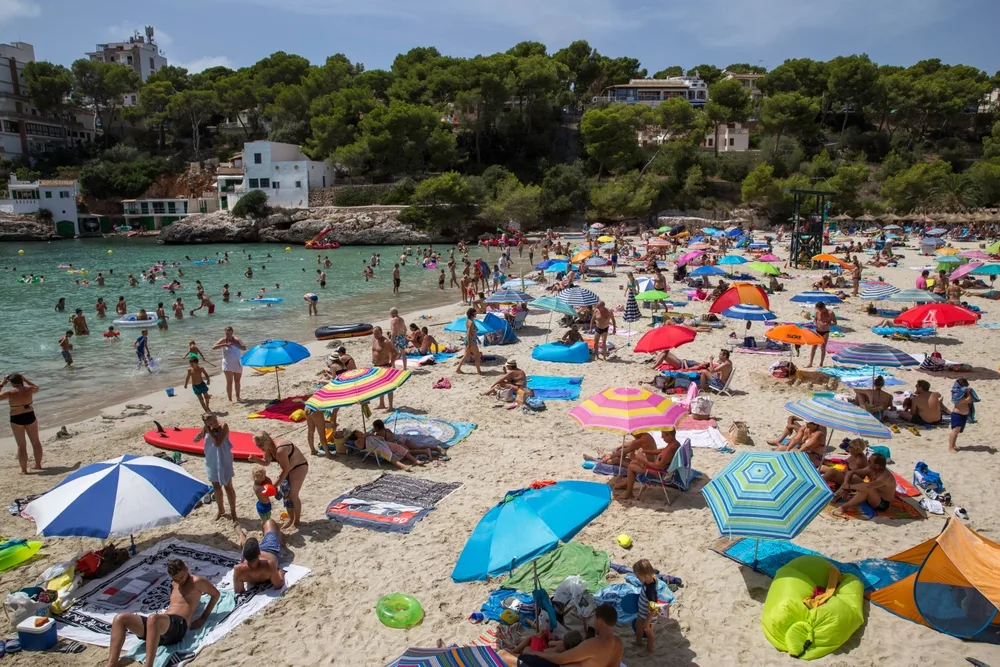 Hundreds of tourists on a beach in Mallorca during the worst time to visit in the summer with teal water still surrounding the beach