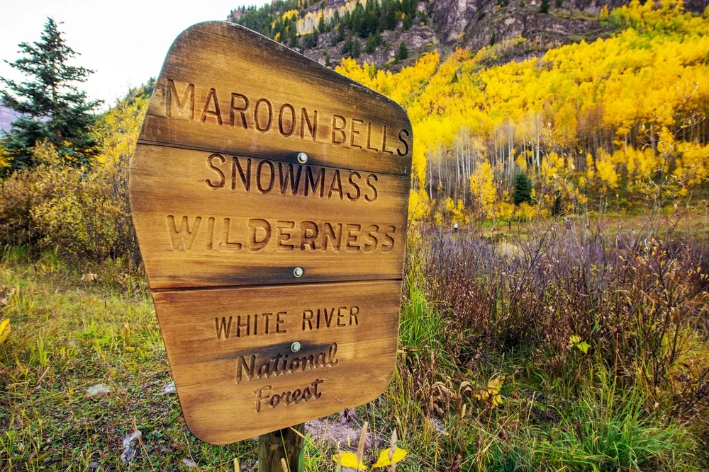 Wooden sign that says Maroon Bells Snowmass pictured during the fall with gorgeous yellow leaves on the trees