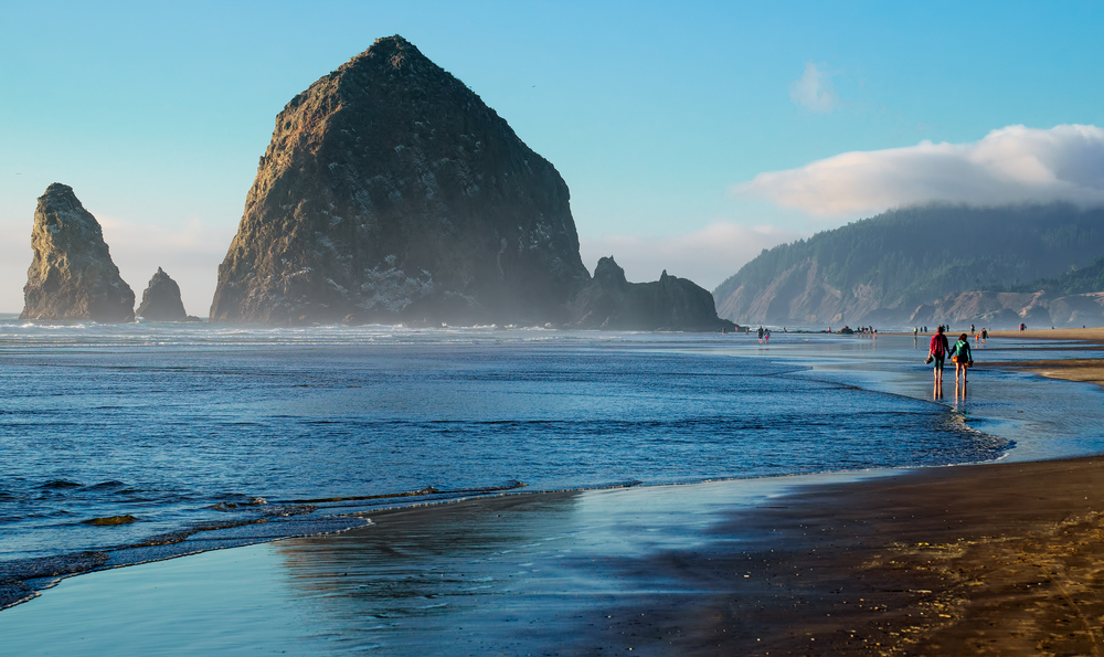 Two people walking along the still water while it laps their feet on the black sand beach pictured during the best time to visit Cannon Beach