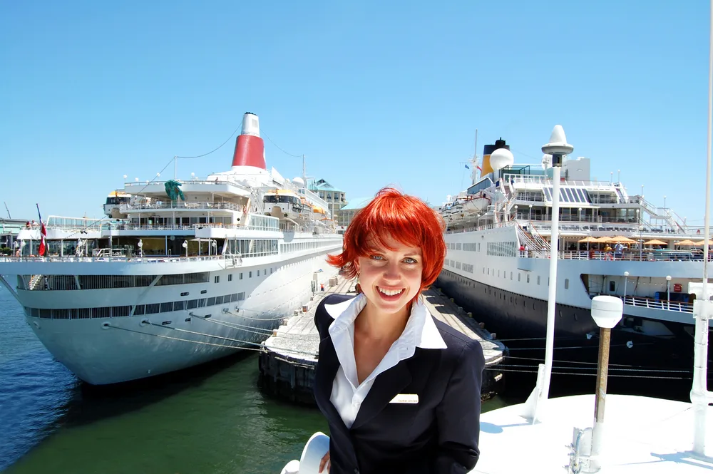 Redhaired woman in a business suit works on a cruise ship standing in front of 2 docked ships for a piece on how to get paid to travel