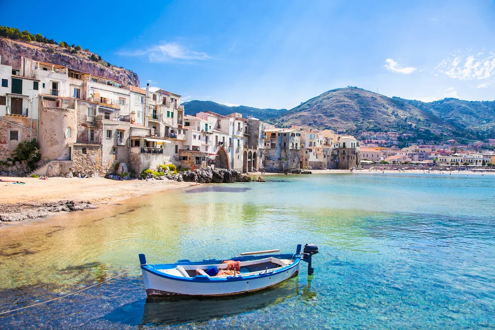 Old wooden blue and white boat pictured on the water that is crystal-clear in front of old buildings overlooking the bay in Cefalu, a must-see destination in Sicily