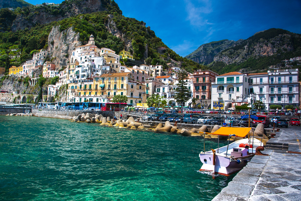 Teal water of Positano as seen from the stone jetty with a boat in the water and the stunning hilltop town with buildings built on the cliffside in the distance