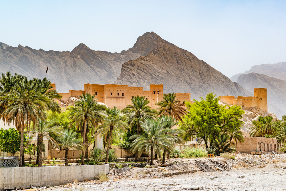 Picturesque view of the Nakhal Fort in Al Batinah with fog rolling over the hills and a clay fort in the foreground with trees surrounding it pictured during the best time to go to Oman