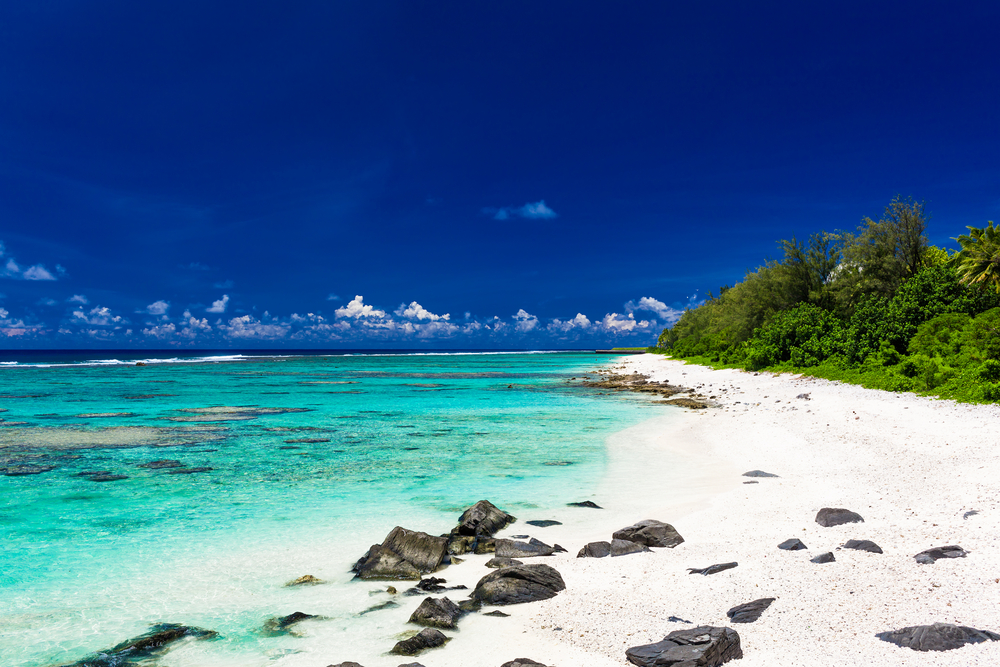 View of a white sand beach on Rarotonga in the Cook Islands with small rocks along the shore and blue skies overhead, a great place to visit in May