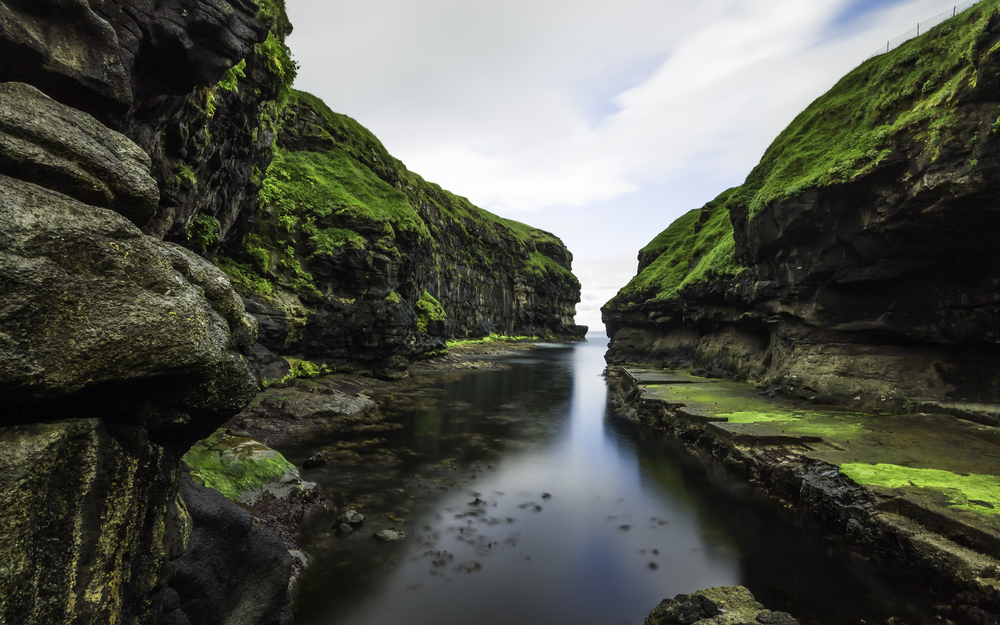 Summer time in the Faroe Islands with a still stream and rocky croppings on either side with moss covering all with cloudy skies above
