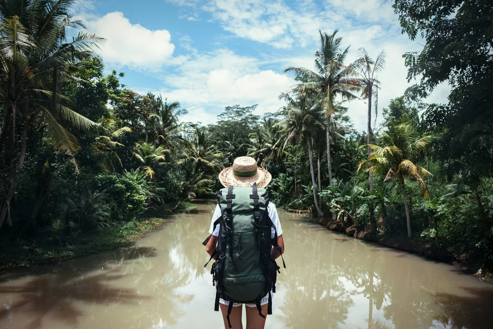 Person traveling wears a hat and stands in front of a muddy river with trees on either side for a piece on how to get paid to travel