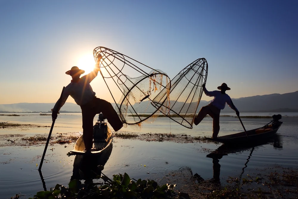 Burmese men in Myanmar pictured doing their one-leg rowing style while holding net in the other hand at dusk