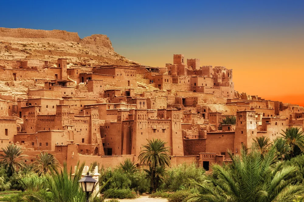Sand-colored buildings and homes lining the cliffs of Ait Ben Haddou, one of the best places to visit in Morocco, pictured at dusk with an orange sky in the background