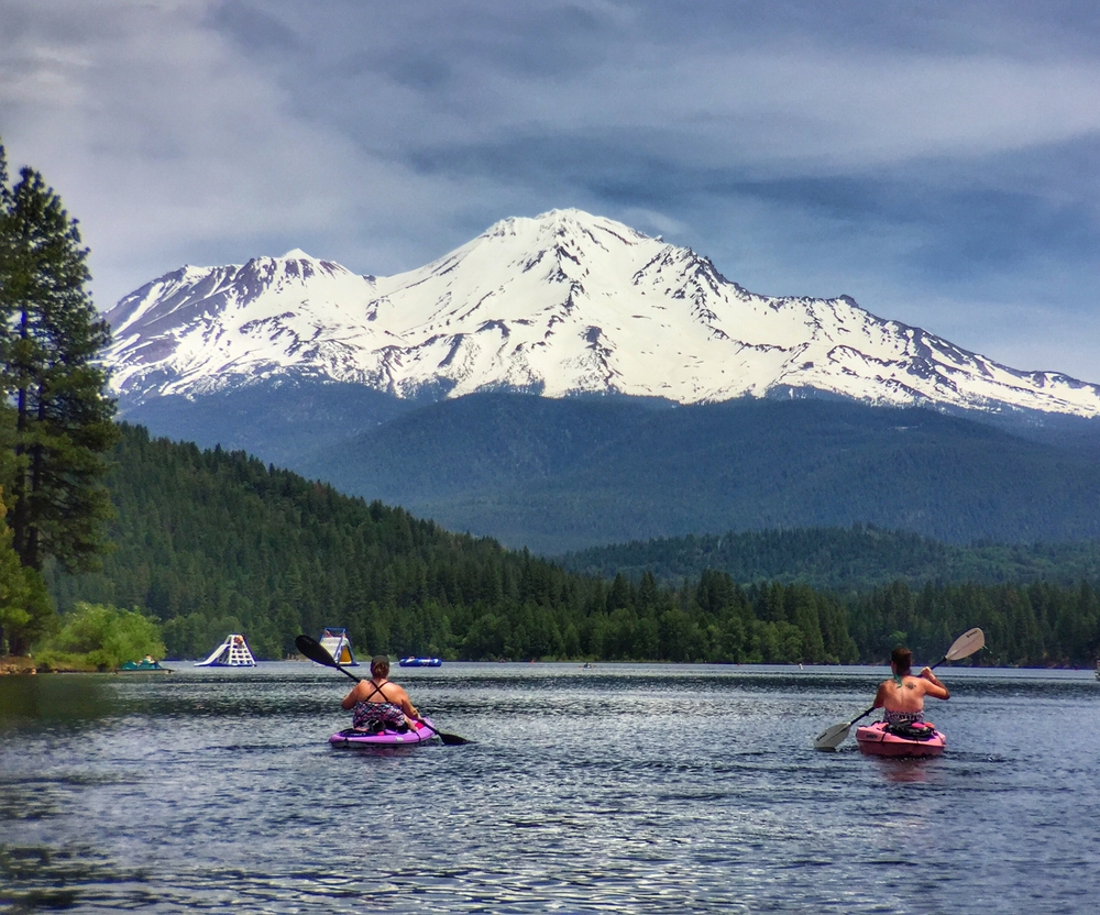 Two young woman paddling on kayaks on a lake below Mount Shasta on a semi-cloudy day