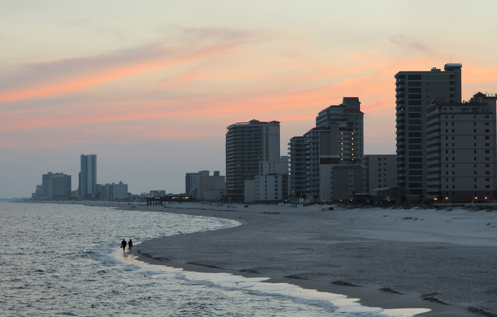 Two people walking alone on the beach in Gulf Shores during the best time to visit while the sun sets over the horizon