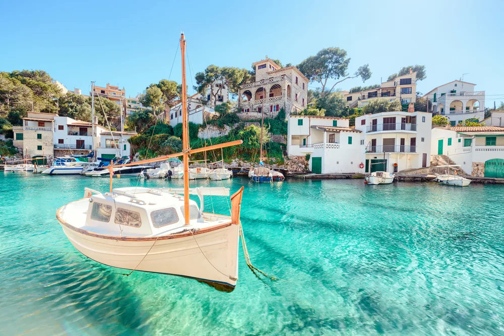 Boat floating on crystal-clear water on the ocean that almost looks fake while buildings surround the scene for a piece on the best time to visit Mallorca