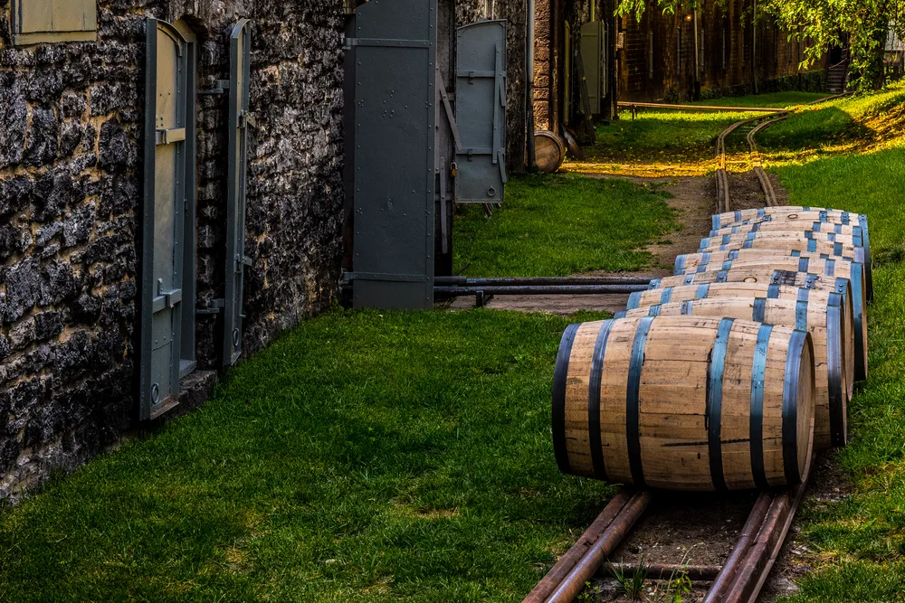 Bourbon barrels pictured rolling along a path outside of a stone wall with metal doors