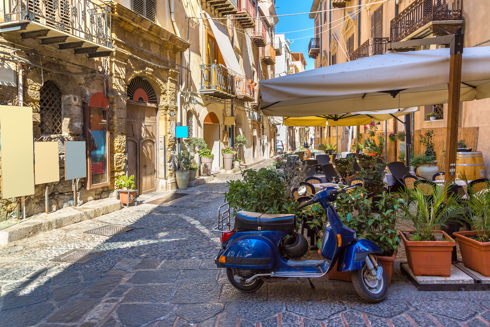 Narrow street in Sicily pictured with a Vespa on its kickstand by flower pots