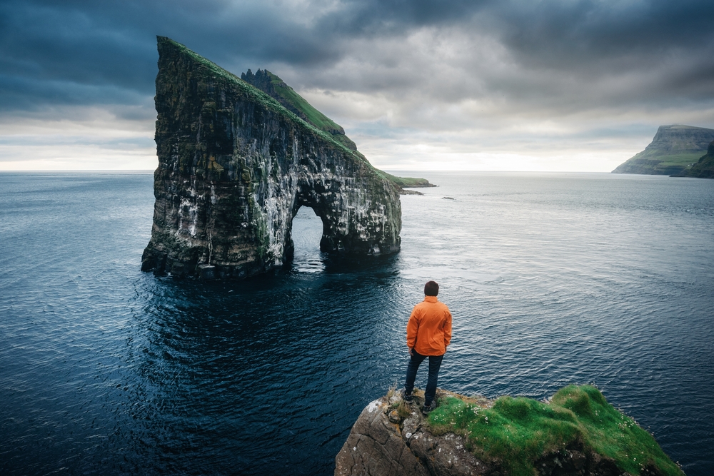 Man standing on the edge of a rock and overlooking the rock with a hole in it (Drangarnir Gates) during the cheapest time to visit the Faroe Islands