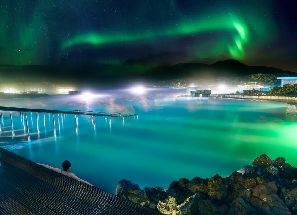 Woman with her arms outstretched pictured below the Northern Lights during the best time to go to the Blue Lagoon in Iceland