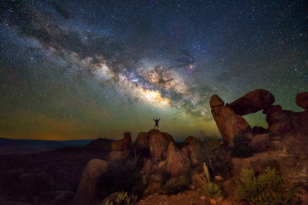 Man standing on a rock with his arms above his head at night in Big Bend National Park, one of the best places to visit in Texas, with a breathtaking view of the sky overhead