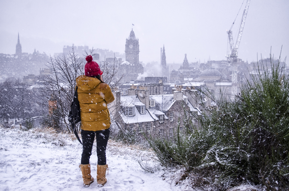 Woman in a red hat and a yellow coat shivering and standing on a hill overlooking Edinburgh during the winter, the worst time to visit