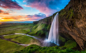 Seljalandsfoss falls pictured pouring into the basin below with water running between the meadows into the distance as seen at sunset