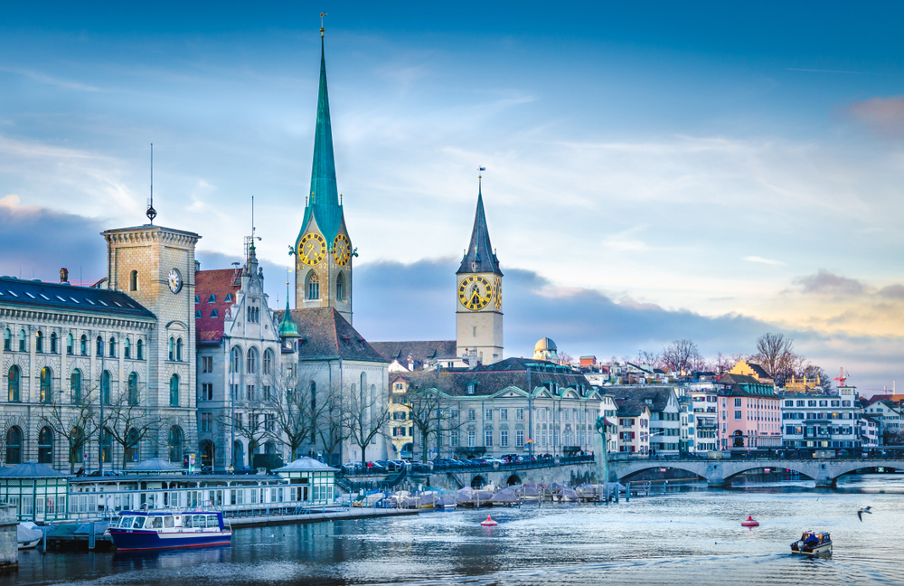 Winter view of Zurich pictured with old buildings with spires and ice on the water for a piece on the best places to visit on a trip to Switzerland