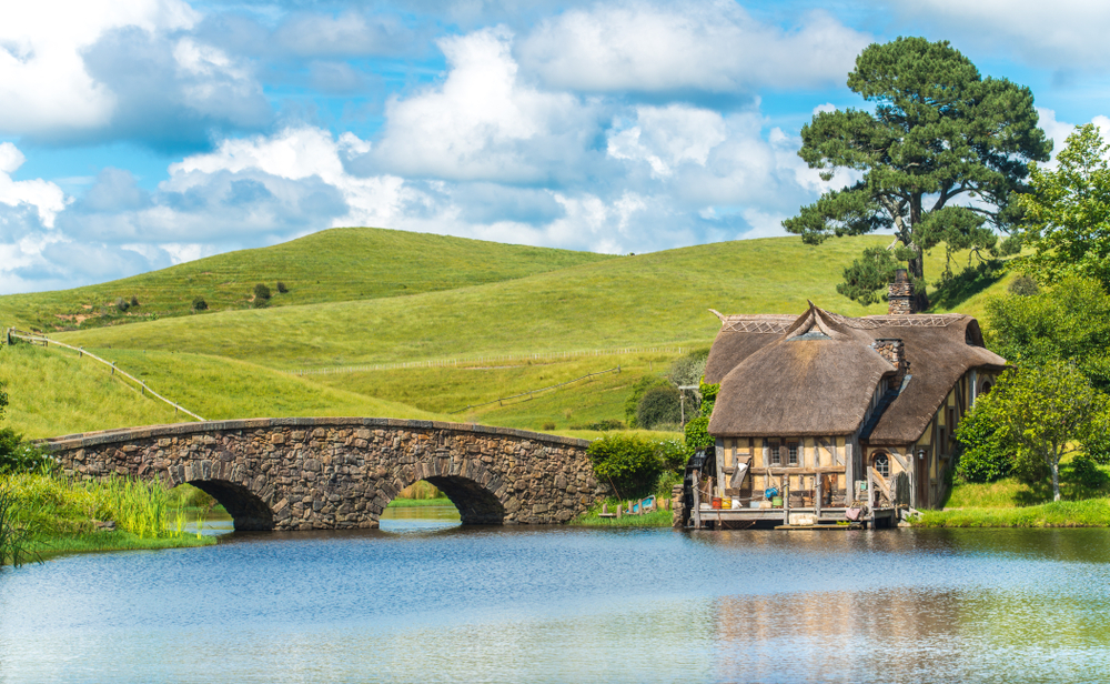 Quaint little town of Matamata, one of the best places to visit in New Zealand, pictured with a stone bridge going over the pond next to a little wooden and thatch roof shack