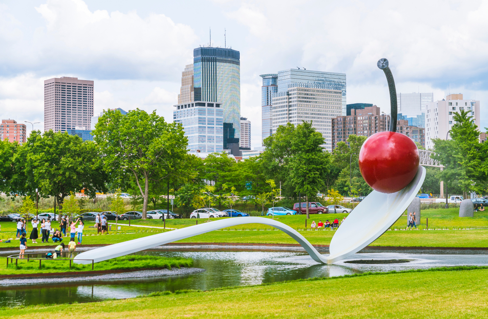 For a piece titled Is Minneapolis Safe to Visit, a giant spoon with a giant cherry on it pictured in a well-manicured park