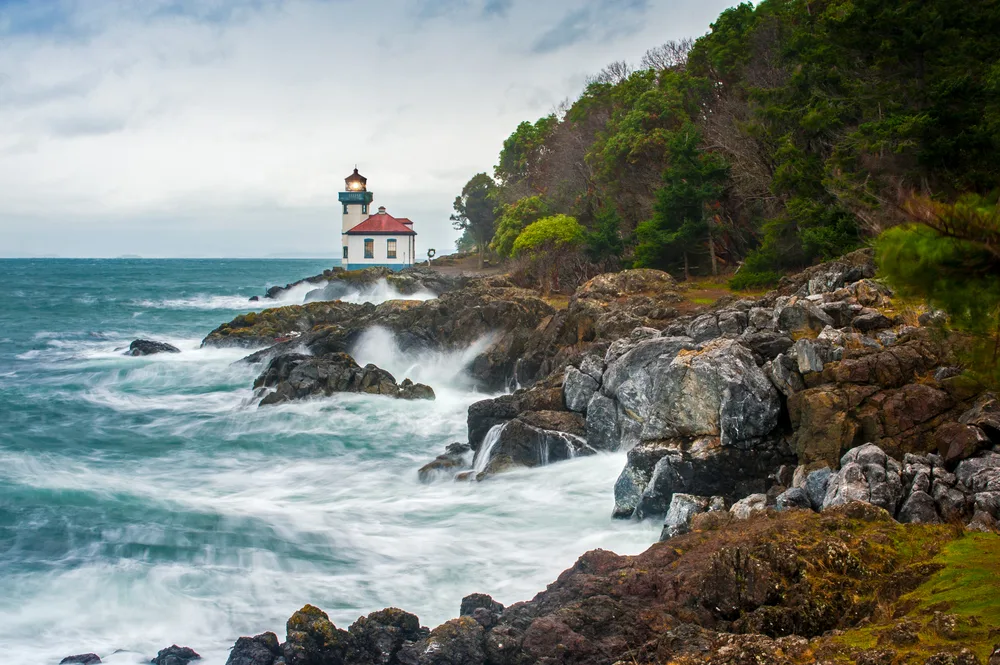 White capped waves crash against the black rocks on the coast of the San Juan Islands with a lighthouse above them