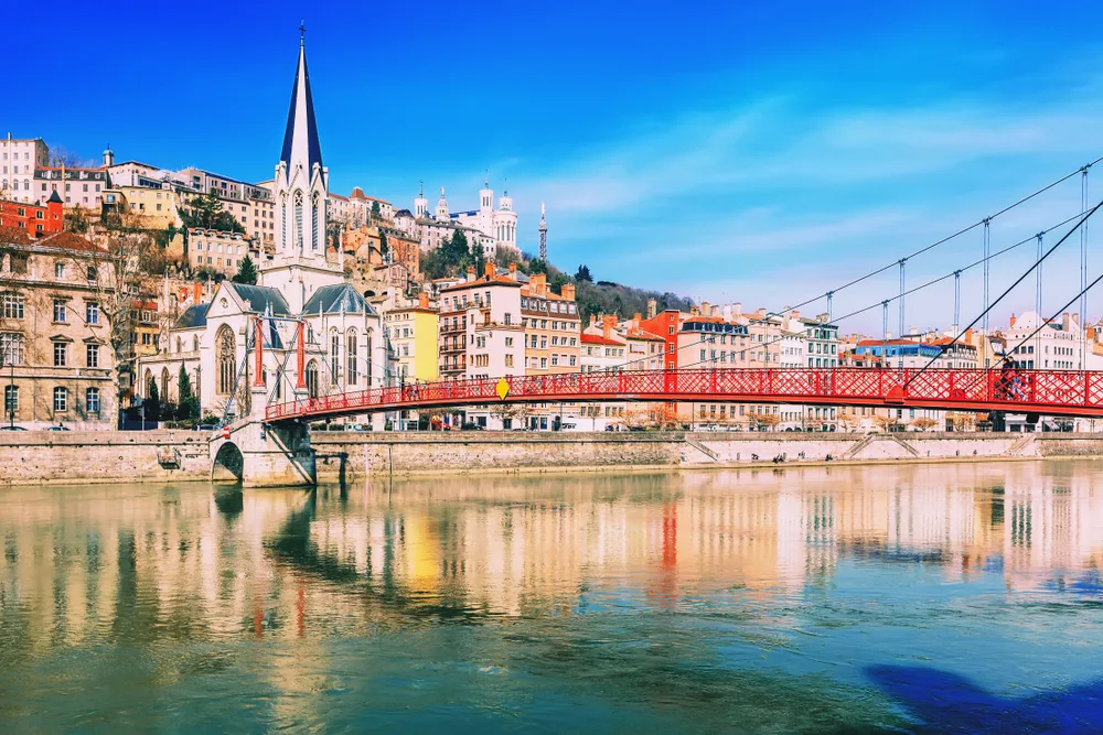 For a suggestion on the best places to visit on a trip to France, a photo of a suspension foot bridge leading to the church on the river of Lyon, as seen on a clear day with old buildings overlooking the river