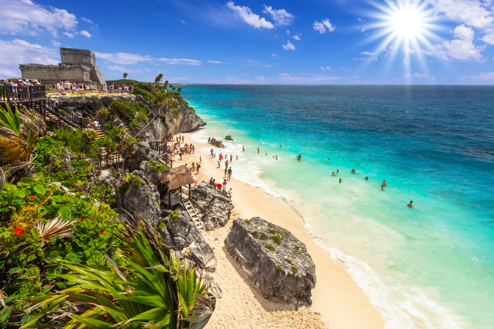 A small number of tourists playing in the water below the ruins of Tulum, a top pick for must-visit places in Central America, and sun shining down from the deep blue sky above