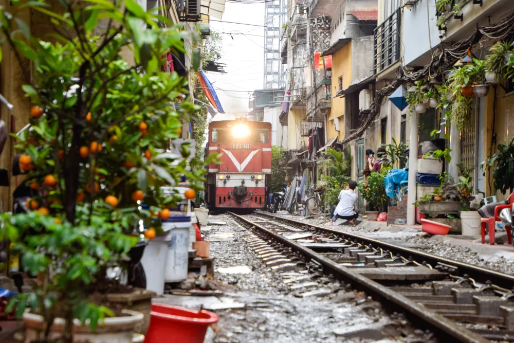 Small red train running through the middle of a bustline city with homes on either side of the tracks, seen at one of Asia's best places to visit, Hanoi