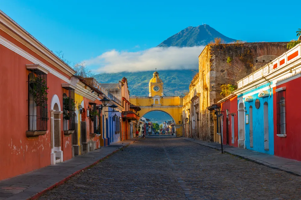 Main road going through colorful red and blue and yellow homes with a yellow arch above the street at the end and the giant volcano in the background in Antigua, a top pick for the best places to visit in Central America