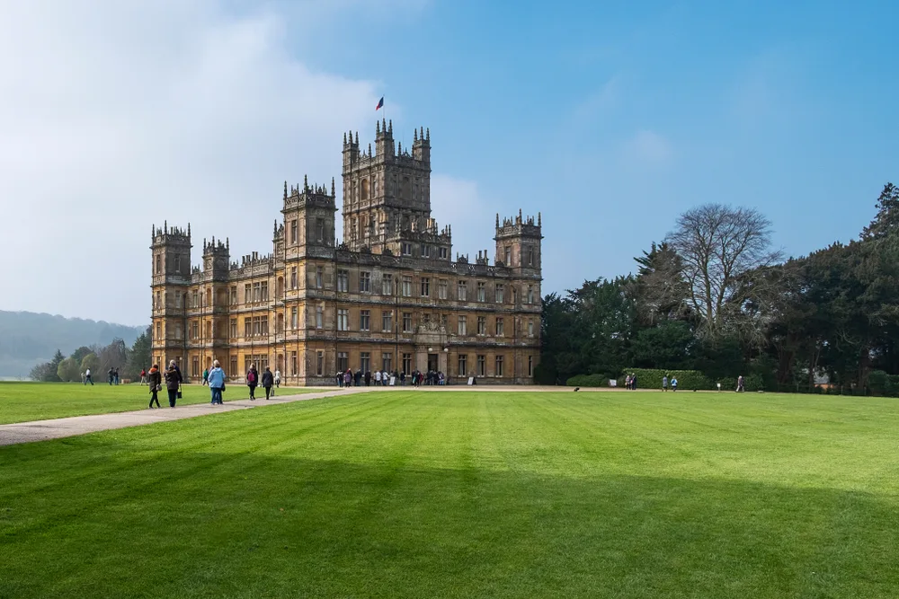 Red brick Highclere Castle pictured on a clear summer's day with green grass in front of the tall trees and a few clouds overhead