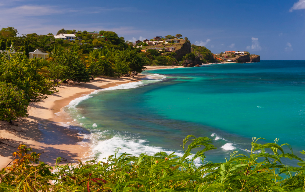 View of Grand Anse Beach in Grenada, one of the best islands in the Caribbean pictured with waves crashing against the sandy shore with blue skies and greenery