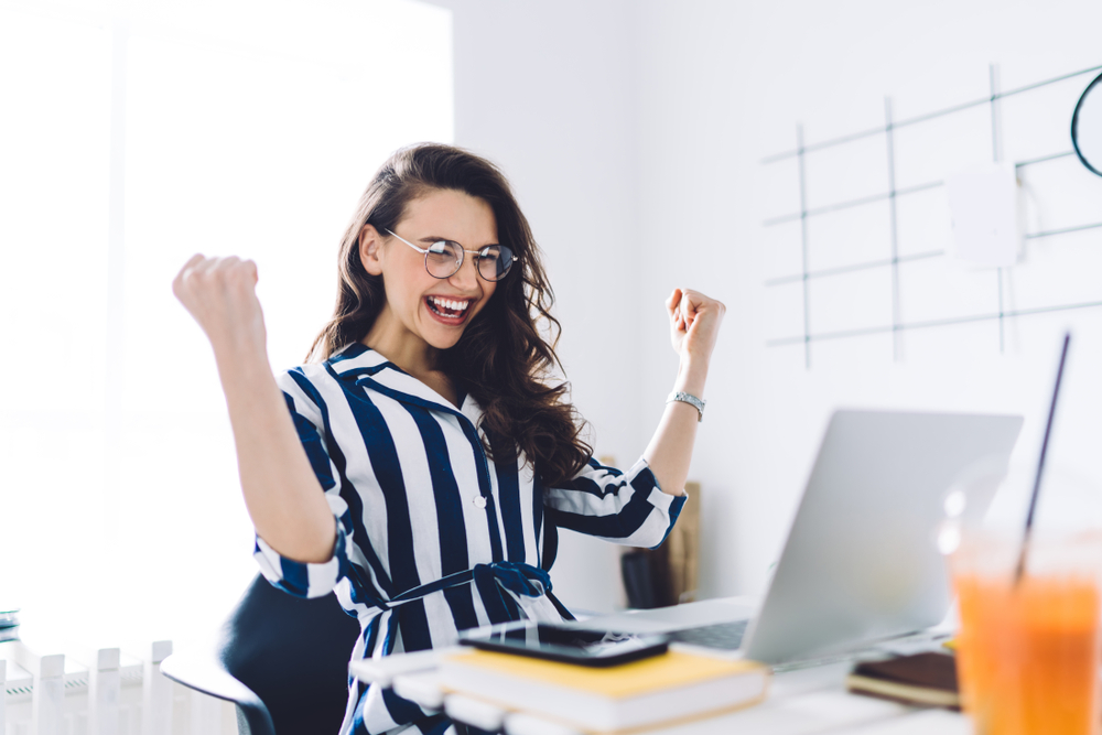 Woman smiles with arms raised at her computer at work to show job satisfaction health benefits of traveling