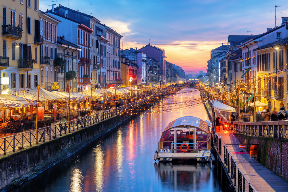Boats on the canal in Navigli at night with restaurants and walkways on either side with lights strung across the water and an orange sky in the background for a piece titled Where to Stay in Milan, Italy