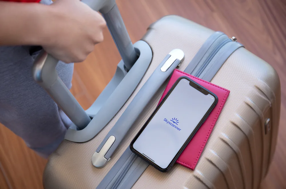 Luggage with smartphone resting on top showing the Skyscanner app screen and passport underneath